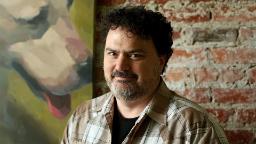تیم شیفر (Tim Schafer) با موهای فر و ریش در مقابل دیوار آجری