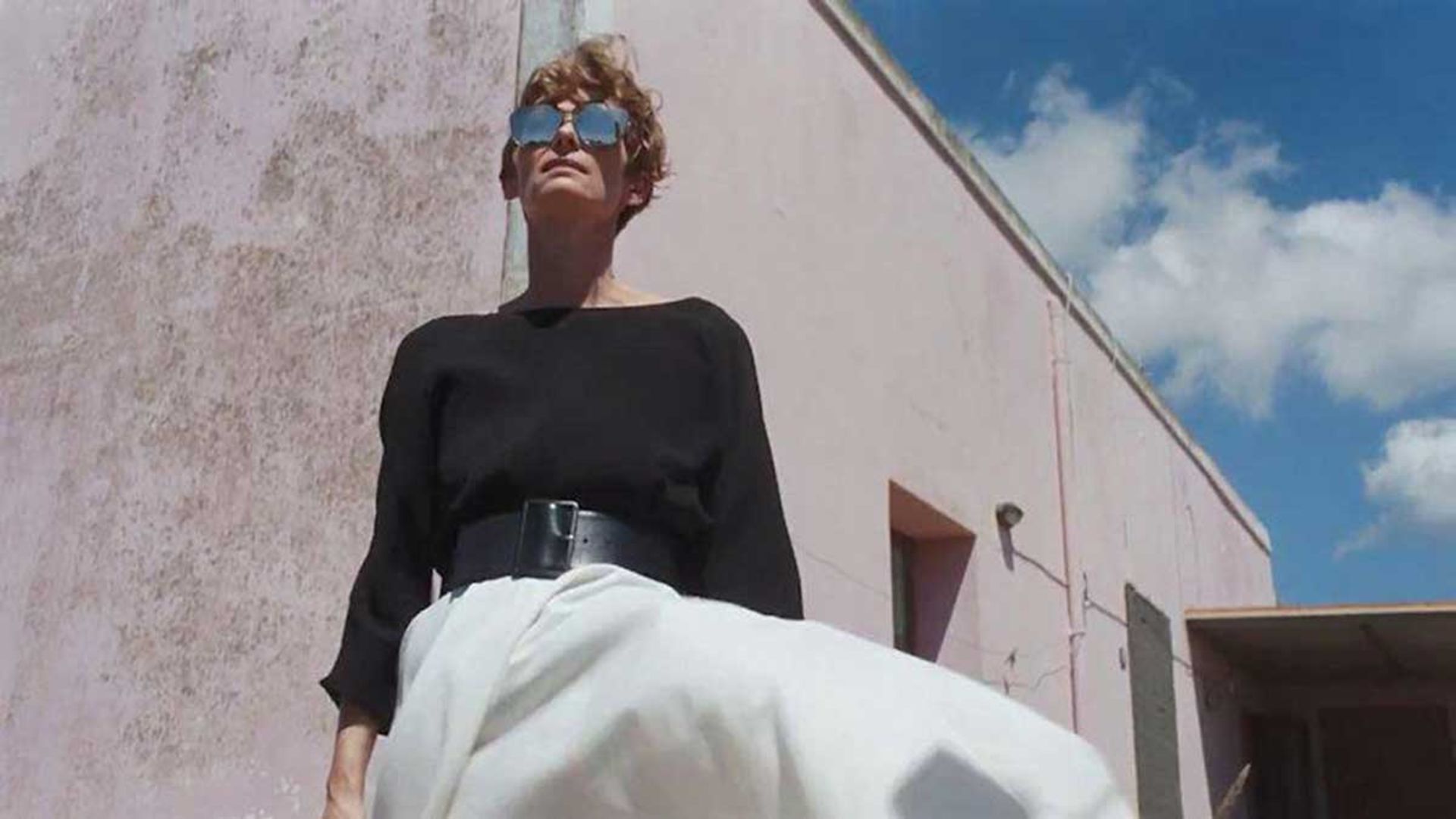 تیلدا سوینتن با دامن سفید، بولیز مشکی و عینک آفتابی در فیلم A Bigger Splash، محصول سال ۲۰۱۵ میلادی