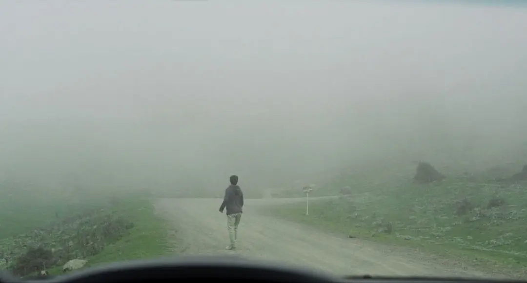 مردی ایستاده در جاده‌ای خاکی در حالی که اطراف‌اش را مه فراگرفته در نمایی از فیلم جاده خاکی به کارگردانی پناه پناهی