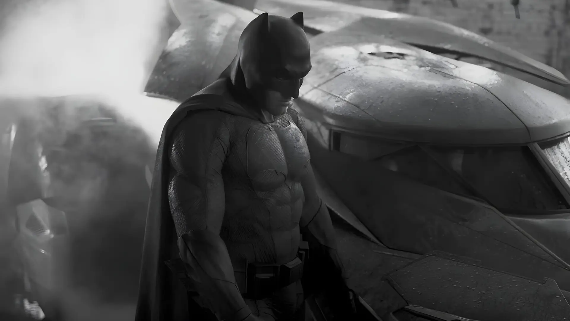 بتمن (Batman) با لباس مخصوص در کنار خودروی بت‌موبیل ایستاده است