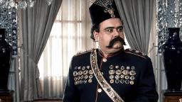 تصویر اکبر عبدی با لباس و کلاه سنتی پادشاهان در یک فضای مجلل در فیلم مظفرنامه