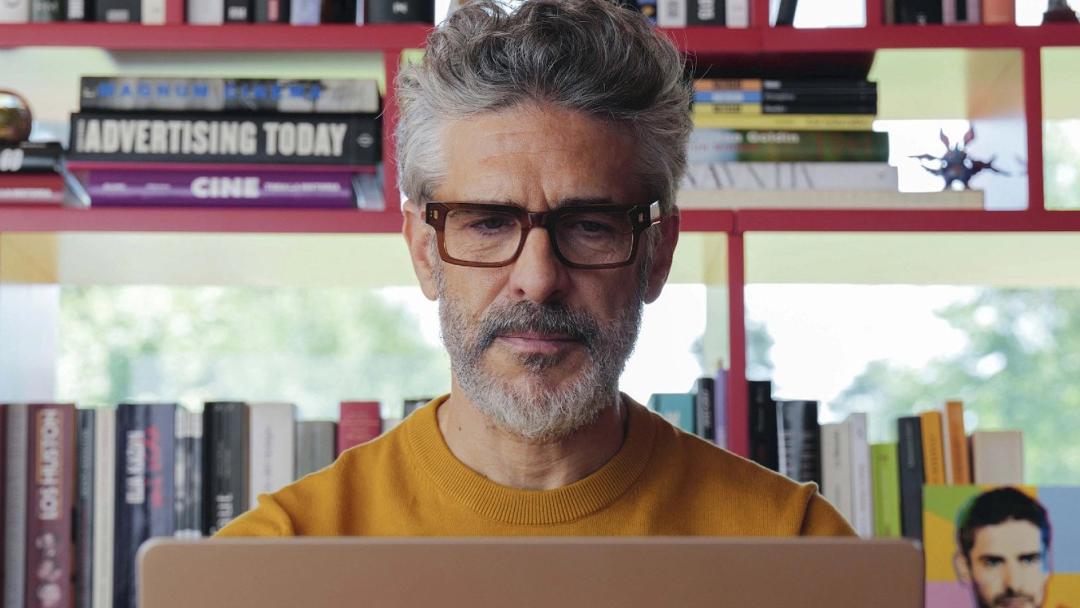 پدرو آلمودوار (Pedro Almodóvar) با عینک در مقابل لپ‌تاپ و کتابخانه